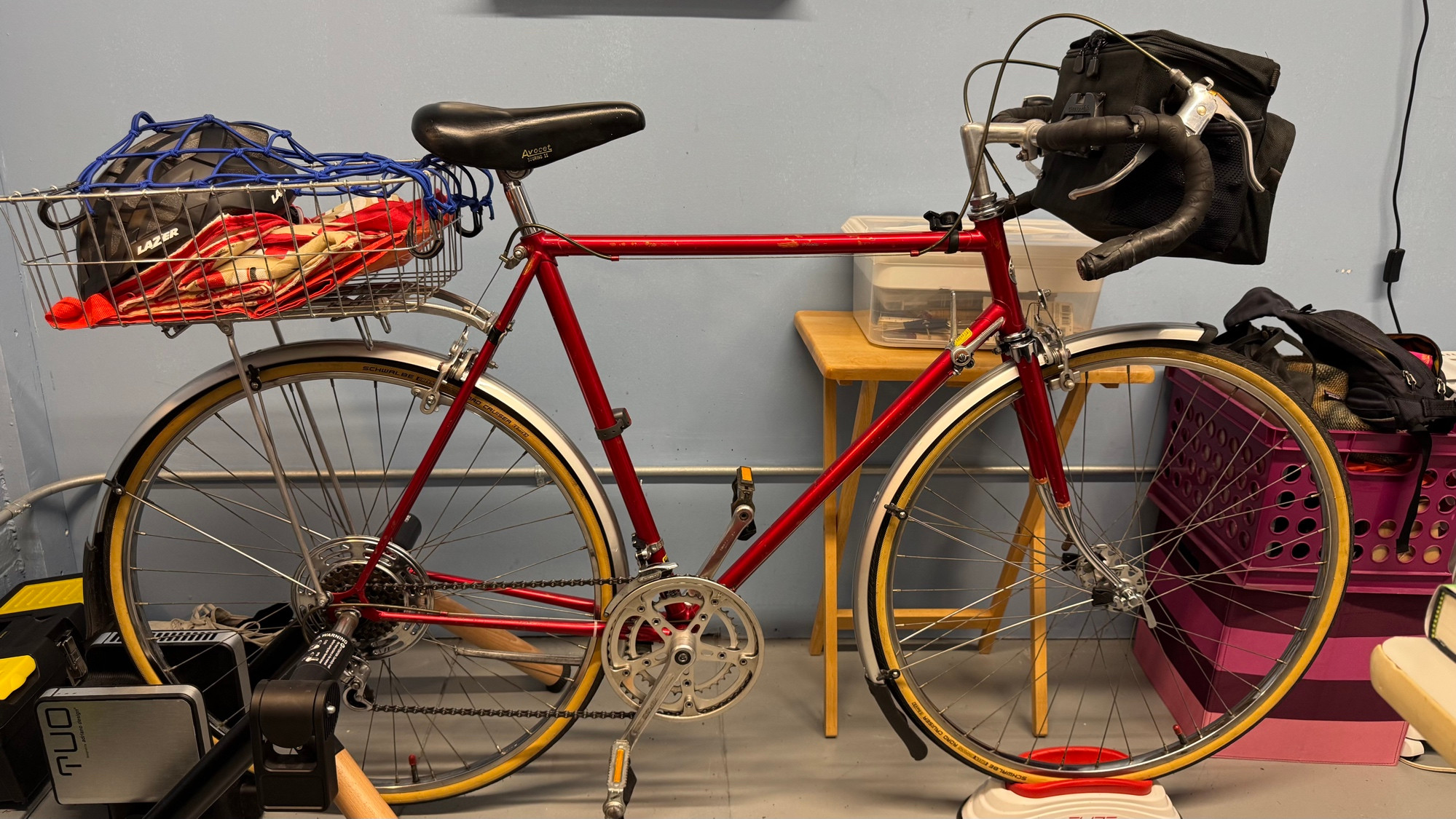 A red Schwinn road bike. On it is attached fenders/mudguards, a handlebar bag, a rear rack, and, on top of the rack, a basket with a blue net to hold stuff in. There are folded shopping totes and a helmet in the basket. The bike is on a stationary trainer that applies friction to the rear wheel.